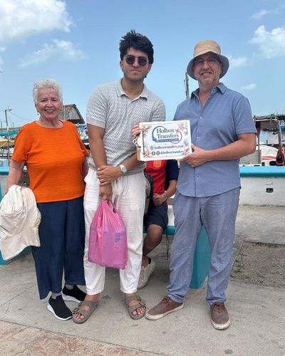 Una familia sostiene el banner de Holbox Transfers en el muelle del ferry, con lanchas atracadas detrás antes de cruzar a Holbox.