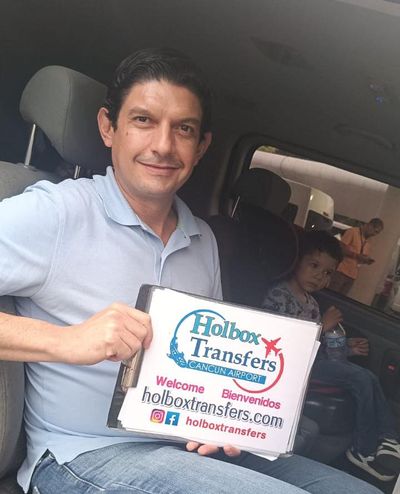 Familia viaja junta en la van rumbo a Holbox, un hombre sonriente sostiene el banner de Holbox Transfers junto a un niño pequeño.
