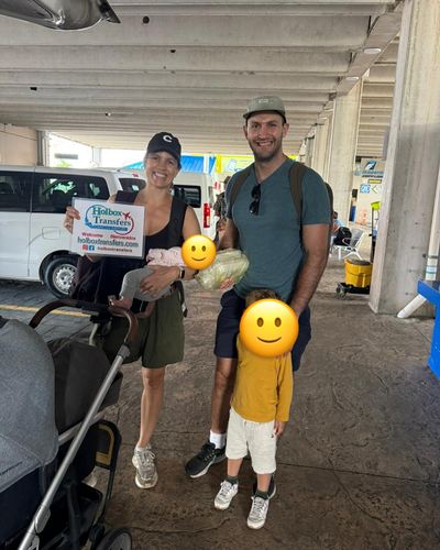 Familia de pie con dos niños junto a la carriola, sosteniendo el banner de Holbox Transfers en el estacionamiento de la terminal del aeropuerto.