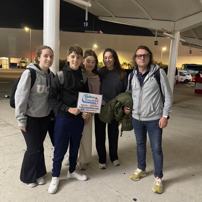 Grupo de amigos sonríe junto al banner de Holbox Transfers en la terminal del aeropuerto durante una llegada nocturna rumbo a Playa del Carmen.