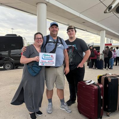 Una familia feliz posa de pie con su equipaje sosteniendo el banner de Holbox Transfers en la terminal del aeropuerto, rumbo a Holbox.