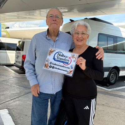 Pareja sonriente abrazada en la terminal, sosteniendo el banner de Holbox Transfers antes de su viaje a Holbox.
