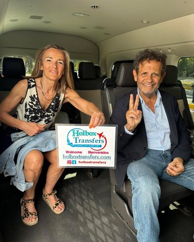 Pareja sonriente hace la señal de amor y paz detrás del banner de Holbox Transfers en la van, en el trayecto Aeropuerto de Cancún - Holbox.