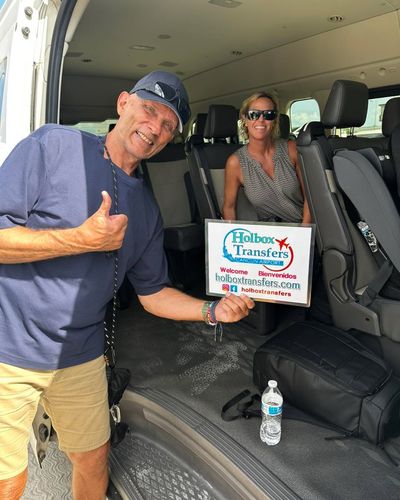 Una pareja sonriente levanta el pulgar sosteniendo el banner de Holbox Transfers dentro de la van antes de partir a Holbox.