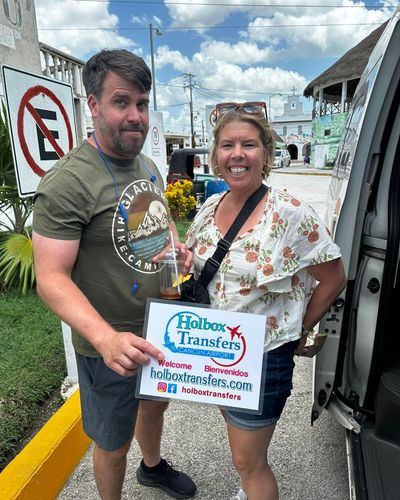 Pareja sonriente sostiene el banner de Holbox Transfers junto a la van durante una parada en el camino a Holbox.