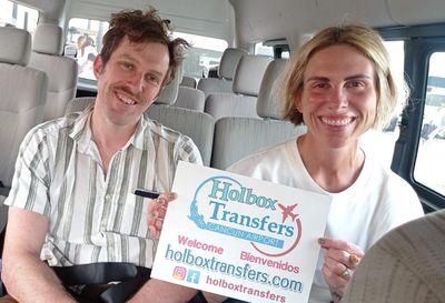 Pareja sonriente sosteniendo el banner de Holbox Transfers dentro de la van, pura sonrisa durante su traslado del Aeropuerto de Cancún a Holbox.