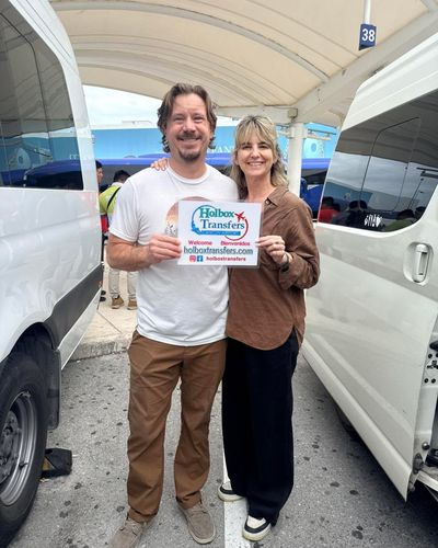 Una pareja sonriente sostiene el banner de Holbox Transfers en la terminal del aeropuerto, recibida para su traslado a Holbox.