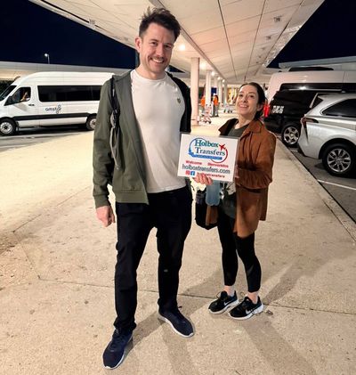 Pareja sonriente sostiene el banner de Holbox Transfers en la terminal del aeropuerto de noche, iniciando su traslado Aeropuerto de Cancún - Holbox.