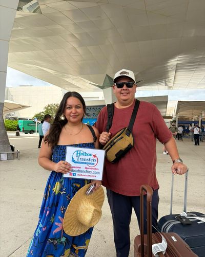 Pareja sonriente sostiene el banner de Holbox Transfers en la terminal del aeropuerto con equipaje, lista para el viaje Aeropuerto de Cancún - Holbox.