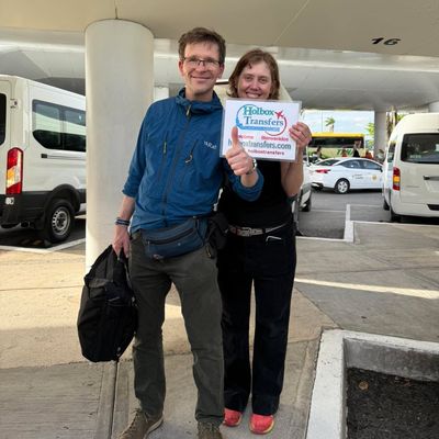 Una pareja sonriente sostiene el banner de Holbox Transfers en la terminal del aeropuerto antes de su traslado a Holbox.