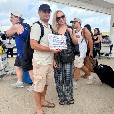 Una pareja sonriente sostiene el banner de Holbox Transfers entre el bullicio de la terminal del aeropuerto antes de salir hacia Holbox.