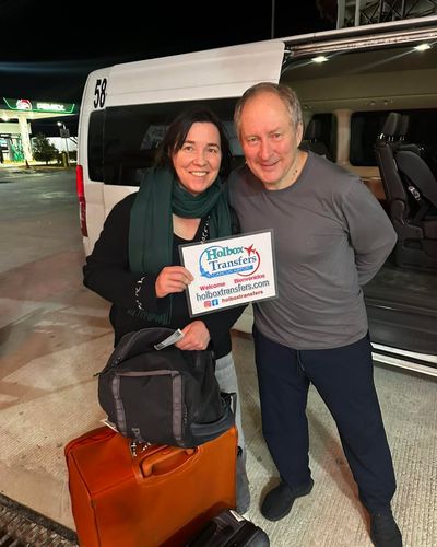 Pareja sonriente sostiene el banner de Holbox Transfers junto a la van de noche con su equipaje, traslado Aeropuerto de Cancún - Playa del Carmen.