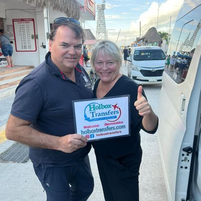 Pareja sonriente sostiene el banner de Holbox Transfers junto a la van en el muelle del ferry de Chiquilá, levantando el pulgar.