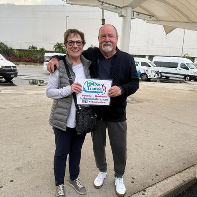 Una pareja sonriente sostiene el banner de Holbox Transfers en la acera del Aeropuerto de Cancún, lista para comenzar su viaje a Holbox.