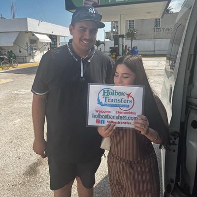 Una pareja sonriente sostiene el banner de Holbox Transfers durante una parada en carretera, continuando su travesía del Aeropuerto de Cancún a Holbox.
