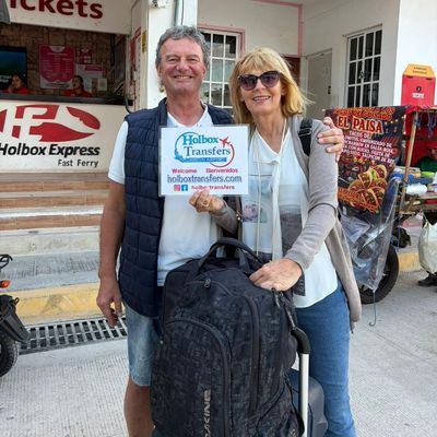 Una pareja sonriente sostiene el banner de Holbox Transfers en la taquilla del ferry, a punto de cruzar a Holbox.