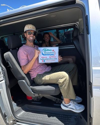 Pareja sonriente sostiene el banner de Holbox Transfers dentro de la van, acomodándose para el trayecto Aeropuerto de Cancún - Holbox.