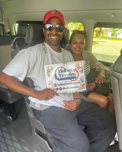 Una pareja sonriente sostiene el banner de Holbox Transfers dentro de la van, con su perrito acompañándolos en el trayecto a Holbox.