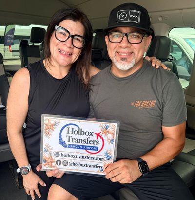 Una pareja sonriente sostiene el banner de Holbox Transfers dentro de la van, puras sonrisas rumbo a Holbox.