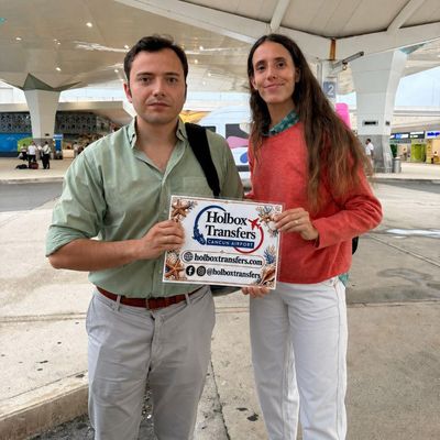 Una pareja sonriente sostiene el banner de Holbox Transfers justo afuera de la terminal del aeropuerto de Cancún antes de comenzar su viaje a Holbox.