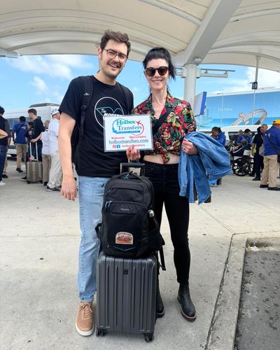 Una pareja sonriente sostiene el banner de Holbox Transfers con su equipaje en la terminal del Aeropuerto de Cancún, lista para Holbox.
