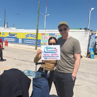Una pareja sonriente sostiene el banner de Holbox Transfers cerca del muelle del ferry, lista para el cruce a Holbox.