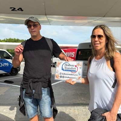 Una pareja sonriente sostiene el banner de Holbox Transfers cerca de la acera de la terminal, lista para comenzar su travesía a Holbox.