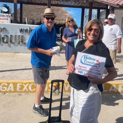 Pareja sonriente sostiene el banner de Holbox Transfers cerca del muelle del ferry, viajeros felices llegando para su travesía a Holbox.