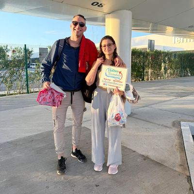 Pareja sonriente sostiene el banner de Holbox Transfers afuera de la terminal con sus maletas, cerrando el traslado Aeropuerto de Cancún - Holbox.