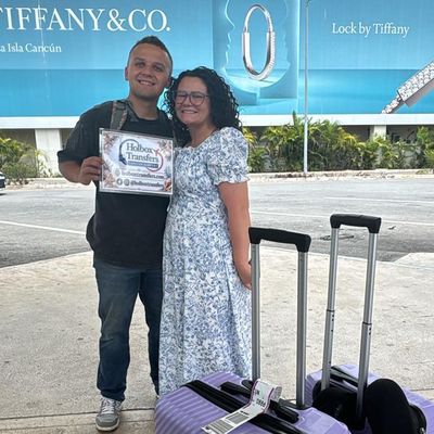 Una pareja sonriente sostiene el banner de Holbox Transfers afuera de la terminal del aeropuerto con equipaje, lista para partir a Holbox.
