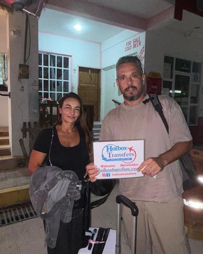 Pareja sonriente sostiene el banner de Holbox Transfers afuera de su alojamiento de noche, recién llegados por la ruta Aeropuerto de Cancún - Holbox.