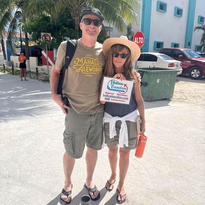 Una pareja sonriente sostiene el banner de Holbox Transfers en una calle de arena en Holbox, ya en su destino.