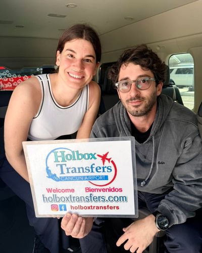 Pareja sonriente sostiene el banner de Holbox Transfers sentada en la van, antes del recorrido Aeropuerto de Cancún - Holbox.