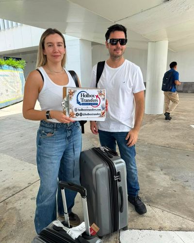Una pareja sonriente sostiene el banner de Holbox Transfers en la terminal, con las maletas listas para el viaje a Holbox.