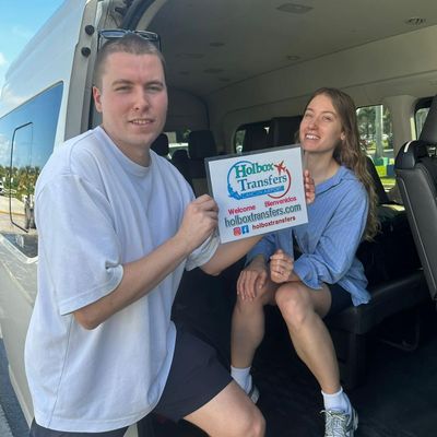 Una pareja sonriente sostiene junta el banner de Holbox Transfers en la puerta de la van, lista para partir a Holbox.