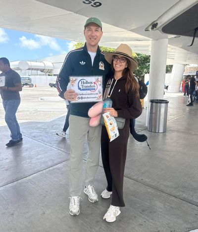 Una pareja sonriente sostiene el banner de Holbox Transfers bajo el techo de la terminal, puras sonrisas antes de emprender el viaje a Holbox.