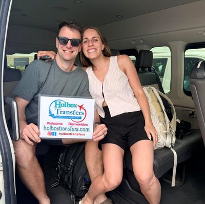 Pareja sonriente se acurruca dentro de la van, sosteniendo junta el banner de Holbox Transfers mientras sonríen para su traslado a Holbox.