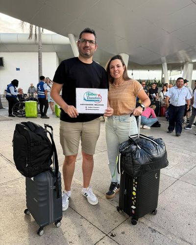 Pareja sonriente posa con su equipaje en la terminal del aeropuerto, sosteniendo el banner de Holbox Transfers antes de su traslado a Holbox.