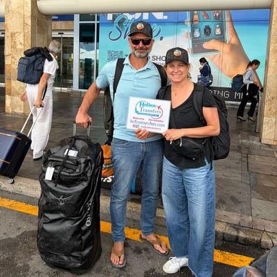 Pareja sonriente posa con equipaje y el banner de Holbox Transfers afuera de la terminal, lista para salir hacia Holbox.