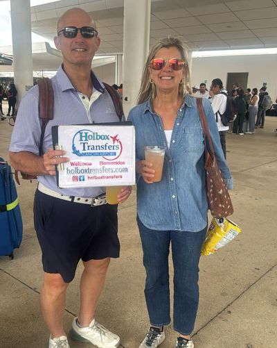 Pareja sonriente posa con bebidas y el banner de Holbox Transfers afuera de la terminal antes del traslado Aeropuerto de Cancún - Holbox.