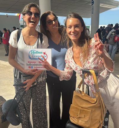 Una pareja sonriente posa sosteniendo el banner de Holbox Transfers junto a la van en el aeropuerto, lista para partir a Holbox.