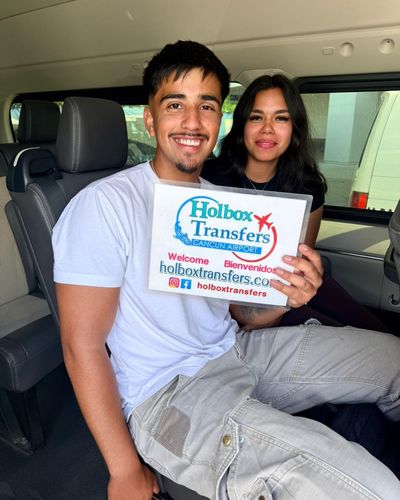 Pareja sonriente posa con el banner de Holbox Transfers dentro de la van, disfrutando el trayecto en el traslado Aeropuerto de Cancún - Holbox.