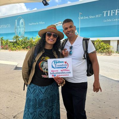 Una pareja sonriente posa con el banner de Holbox Transfers afuera de la terminal del aeropuerto, pulgares arriba antes de partir a Holbox.
