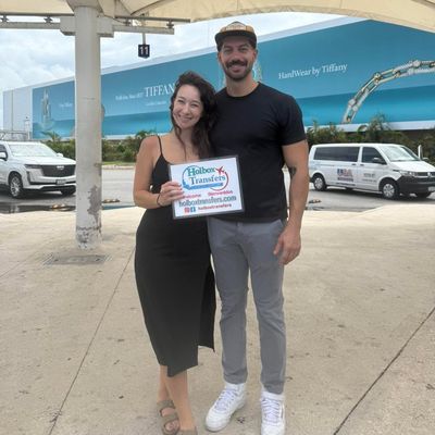 Pareja sonriente posa con el banner de Holbox Transfers afuera de la terminal del aeropuerto, lista para comenzar su viaje a Holbox.