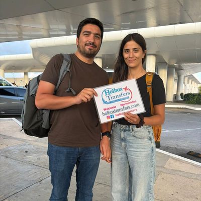 Una pareja sonriente posa con el banner de Holbox Transfers bajo el techo de la terminal en su camino a Holbox.