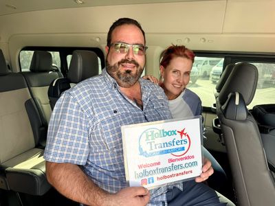 Una pareja sonriente posa dentro de la van, sosteniendo el banner de Holbox Transfers, iniciando su trayecto del Aeropuerto de Cancún a Holbox.