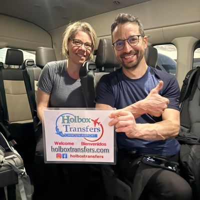 Pareja sonriente posa dentro de la van con el pulgar arriba, sosteniendo el banner de Holbox Transfers antes de su viaje a Holbox.