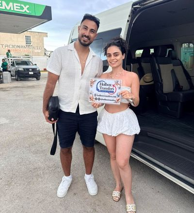 Pareja sonriente posando junto a la van con el banner de Holbox Transfers durante una parada de descanso en el camino a Holbox.