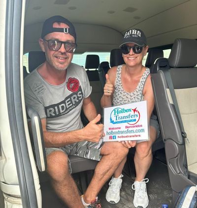Pareja sonriente sentada dentro de la van levanta el pulgar mientras sostiene el banner de Holbox Transfers rumbo a Holbox.