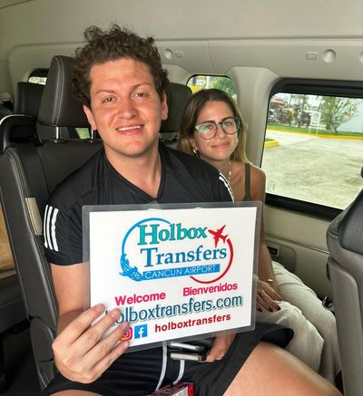 Una pareja sonriente sentada dentro de la van sostiene el banner de Holbox Transfers en su camino a Holbox.